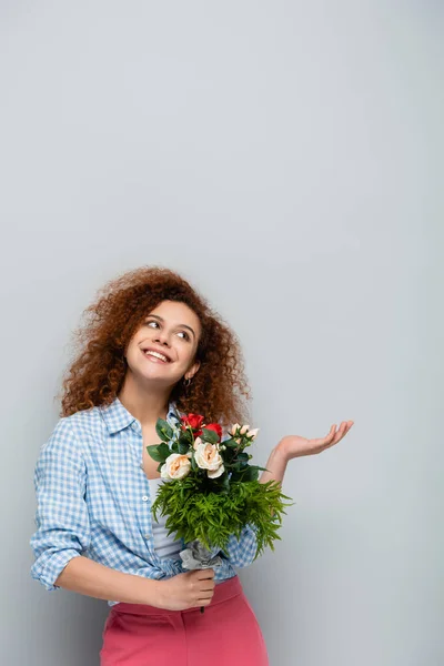 Mulher alegre olhando para cima e apontando com a mão enquanto segurando flores no fundo cinza — Fotografia de Stock