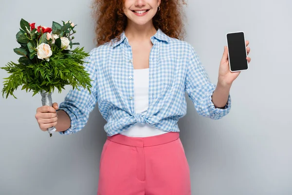 Vista recortada de la mujer sonriente sosteniendo flores y teléfono inteligente con pantalla en blanco sobre fondo gris - foto de stock