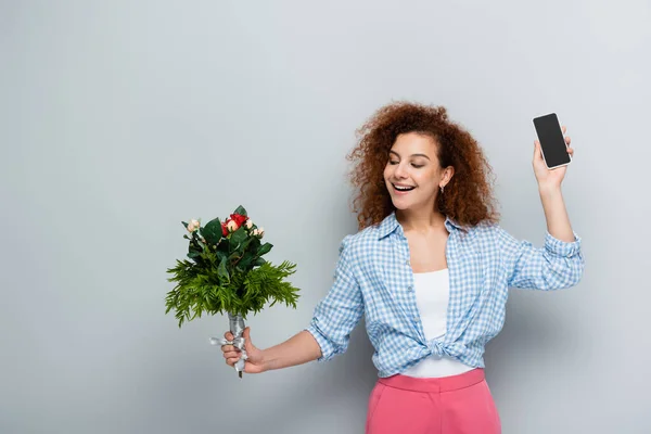 Felice donna che mostra smartphone con schermo bianco mentre tiene i fiori su sfondo grigio — Foto stock