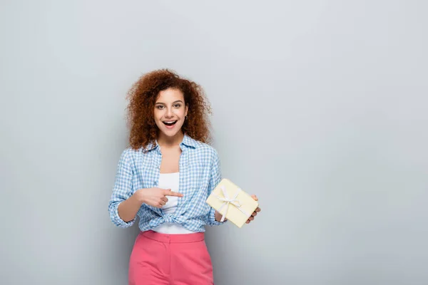 Cheerful woman pointing at gift box while looking at camera on grey background — Stock Photo