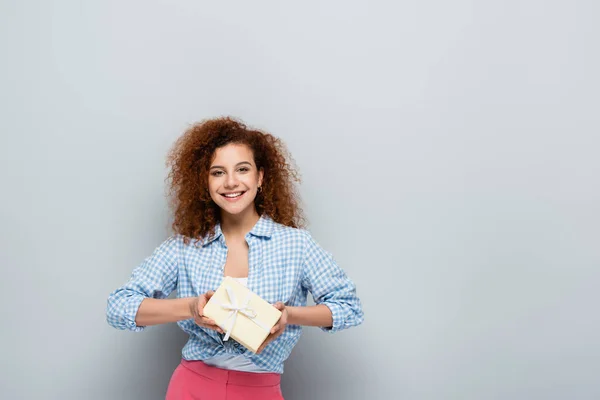 Glückliche Frau blickt in die Kamera, während sie Geschenk auf grauem Hintergrund hält — Stockfoto