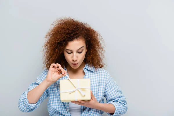 Любопытная женщина в клетчатой рубашке, открывающая подарочную коробку, изолированную на сером — стоковое фото