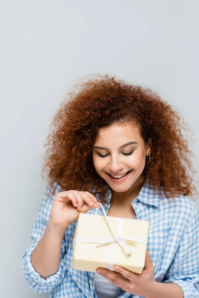 Zufriedene Frau mit welligem Haar öffnet Geschenkbox isoliert auf grau — Stockfoto