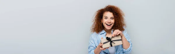 Erstaunte Frau blickt in die Kamera, während sie das Geschenk öffnet, isoliert auf einem grauen Banner — Stockfoto