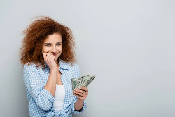 Радостная женщина с рукой рядом с лицом держа доллары на сером фоне — стоковое фото