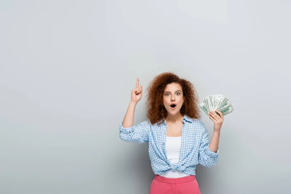 Excited woman pointing up with finger while holding dollars on grey background — Stock Photo
