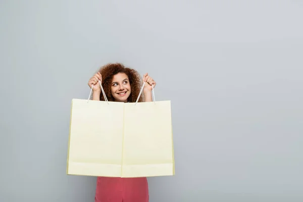 Glückliche, lockige Frau, die wegschaut, während sie Einkaufstüten isoliert auf grau hält — Stockfoto