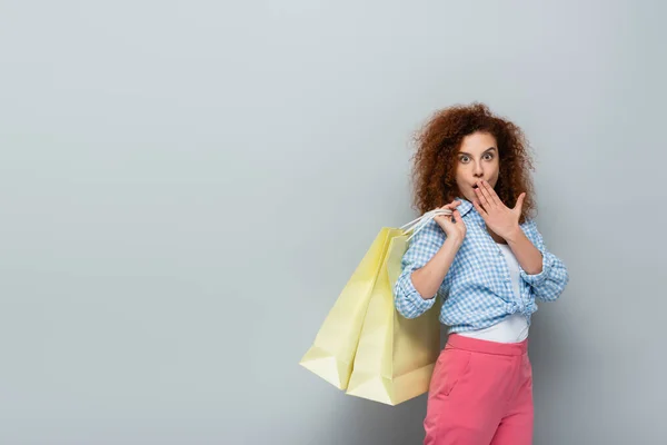 Mulher surpreso cobrindo boca com a mão enquanto segurando sacos de compras em fundo cinza — Fotografia de Stock