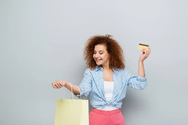 Lockige Frau lächelt, während sie Kreditkarte und Einkaufstasche auf grauem Hintergrund hält — Stockfoto
