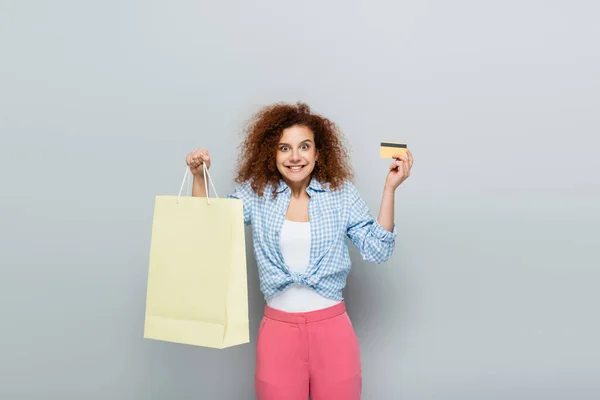 Excited woman with curly hair holding credit card and shopping bag on grey background — Stock Photo