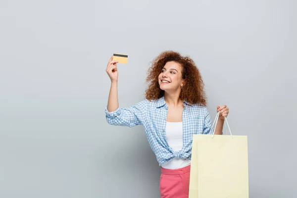 Mulher alegre em camisa xadrez segurando cartão de crédito e saco de compras em fundo cinza — Fotografia de Stock