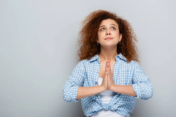 Кудрявая женщина с молитвенными руками смотрит вверх на сером фоне — стоковое фото
