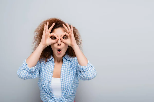 Femme excitée avec des cheveux bouclés imitant les lunettes avec les mains sur fond gris — Photo de stock