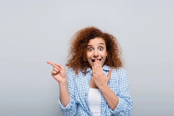 Schockierte Frau zeigt mit dem Finger und bedeckt Mund mit Hand isoliert auf grau — Stockfoto