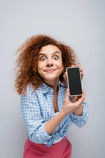 Mujer emocionada haciendo muecas mientras muestra teléfono inteligente con pantalla en blanco aislado en gris - foto de stock