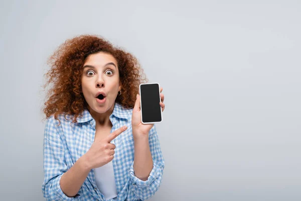 Femme étonnée regardant caméra tout en pointant vers le téléphone portable avec écran vide isolé sur gris — Photo de stock