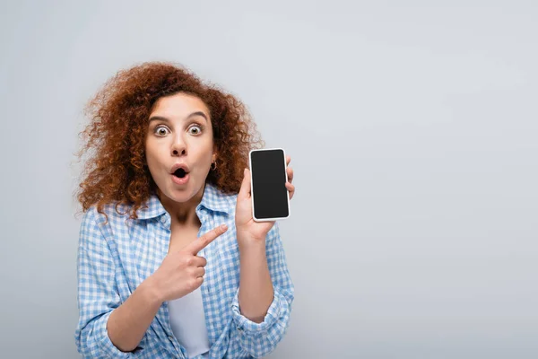 Donna stupita guardando la fotocamera mentre indicava il cellulare con schermo vuoto isolato sul grigio — Foto stock