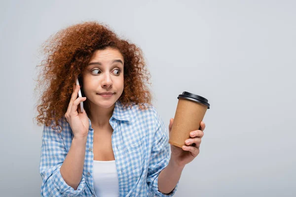 Confused woman looking at paper cup while talking on mobile phone isolated on grey — Stock Photo