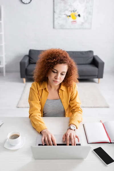 Freelancer com cabelo ondulado digitando no laptop perto do smartphone com tela em branco e xícara de café — Fotografia de Stock