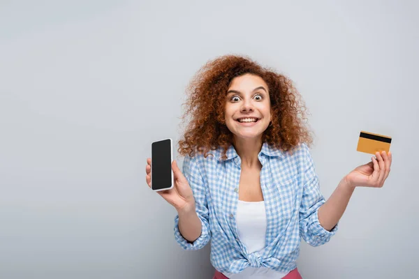 Verblüffte Frau mit Kreditkarte und Handy mit leerem Bildschirm auf grauem Hintergrund — Stockfoto