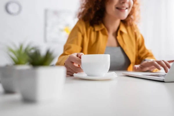 Selective focus of coffee cup near smiling freelancer on blurred background, cropped view — Stock Photo