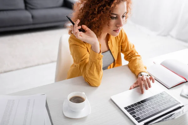 Junge Freiberuflerin mit welligem Haar hält Stift in der Hand und tippt zu Hause am Laptop — Stockfoto