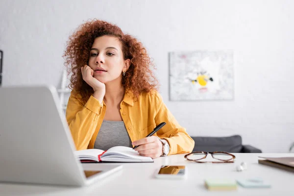 Joven freelancer con el pelo ondulado que sostiene la pluma cerca del cuaderno y del ordenador portátil en primer plano borroso - foto de stock