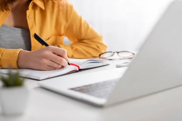 Cropped view of freelancer writing in notebook near laptop on blurred foreground — Stock Photo