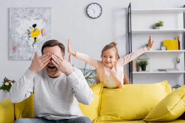 Padre positivo cubriendo los ojos cerca de la hija en la sala de estar - foto de stock