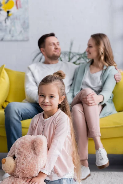 Smiling kid holding teddy bear near parents on blurred background — Stock Photo