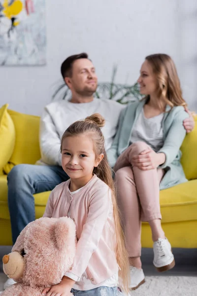 Lächelndes Kind hält Teddybär in der Nähe der Eltern auf verschwommenem Hintergrund — Stockfoto