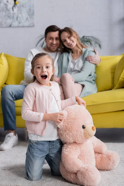 Niño sorprendido sosteniendo oso de peluche cerca de los padres en el sofá sobre fondo borroso - foto de stock