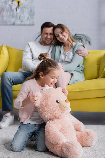 Niño mirando oso de peluche cerca de los padres borrosos en la sala de estar - foto de stock