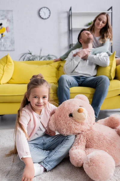 Fille avec ours en peluche assis près des parents flous à la maison — Photo de stock