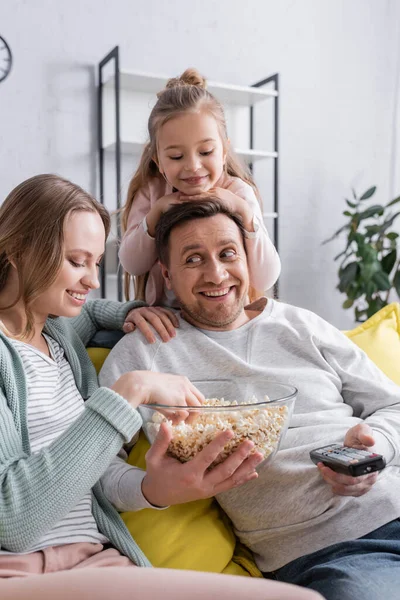 Homme souriant avec pop-corn et télécommande assis près de la femme et de l'enfant — Photo de stock