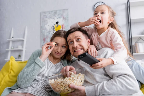 Scared man holding popcorn near wife and child with remote controller — Stock Photo