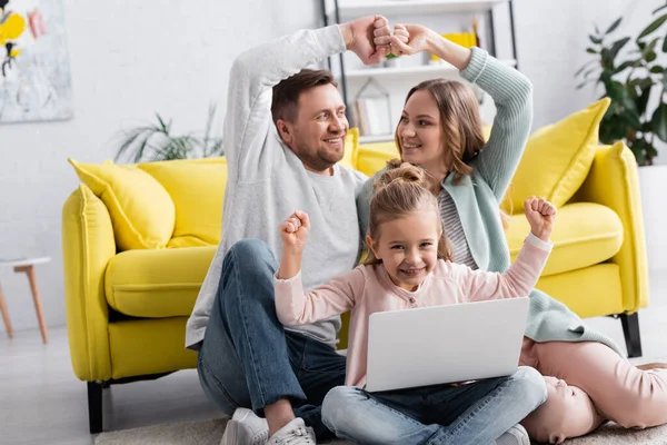 Chica alegre con portátil que muestra sí gesto cerca de los padres en el suelo en la sala de estar - foto de stock