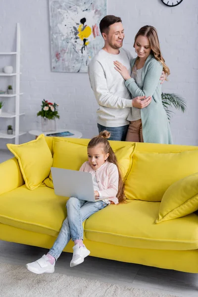 Amazed kid using laptop near cheerful parents hugging at home — Stock Photo