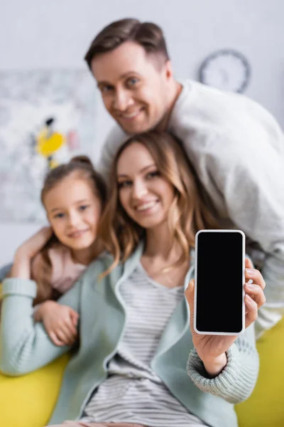 Smartphone con pantalla en blanco en la mano de la mujer sonriente cerca de la familia sobre fondo borroso — Stock Photo
