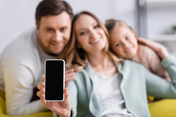 Mujer en fondo borroso sosteniendo teléfono inteligente con pantalla en blanco cerca de la familia - foto de stock