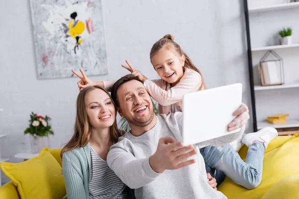Smiling child showing bunny ears with fingers near parents taking selfie on digital tablet — Stock Photo