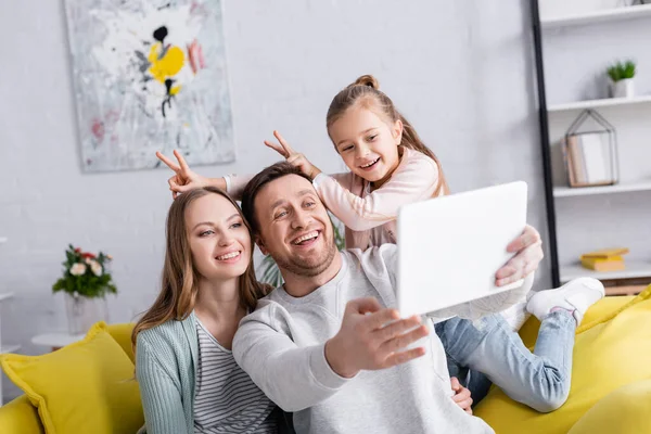 Lächelndes Kind zeigt Hasenohren mit Fingern in der Nähe von Eltern, die Selfie auf digitalem Tablet machen — Stockfoto