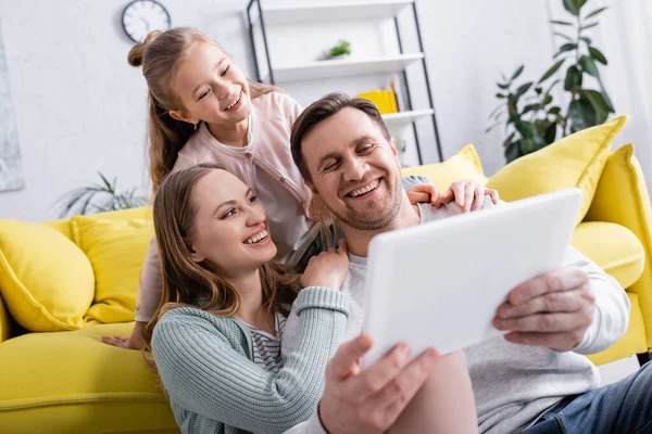 Woman hugging husband with digital tablet on blurred foreground near kid on couch — Stock Photo