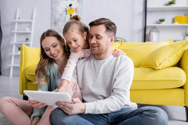 Niño abrazando padre con tableta digital cerca de la madre sonriente en la sala de estar - foto de stock