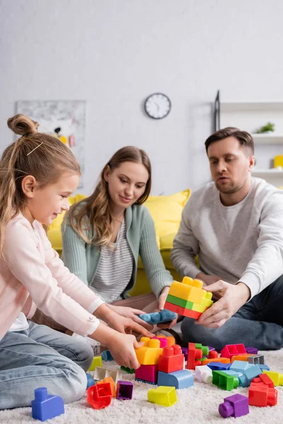 Kid playing building blocks on carpet near parents on blurred background — Stock Photo