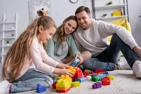 Sonriente niño jugando bloques de construcción en la alfombra cerca de los padres borrosos - foto de stock