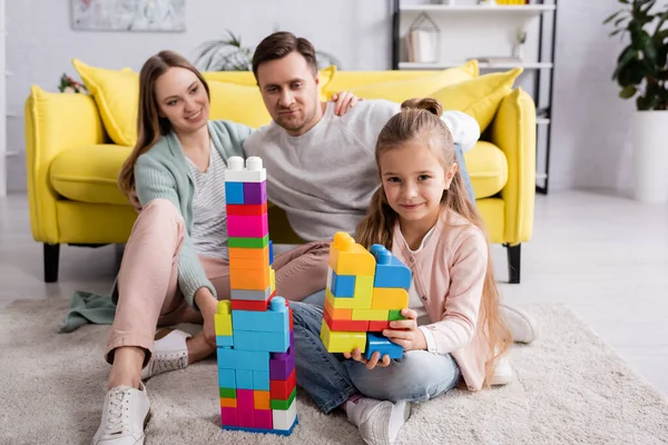 Enfant avec des blocs de construction regardant la caméra tandis que les parents embrassent à la maison — Photo de stock