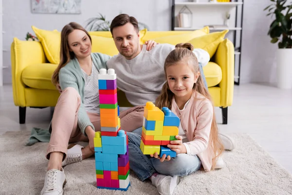 Ragazzo con blocchi di costruzione guardando la fotocamera mentre i genitori abbracciano a casa — Foto stock