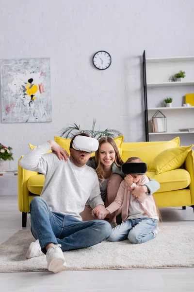 Femme souriante assise près du mari et de la fille dans des casques vr sur le tapis — Photo de stock