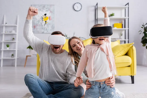 Muchacha y padre emocionados usando auriculares de realidad virtual cerca de la madre en casa - foto de stock